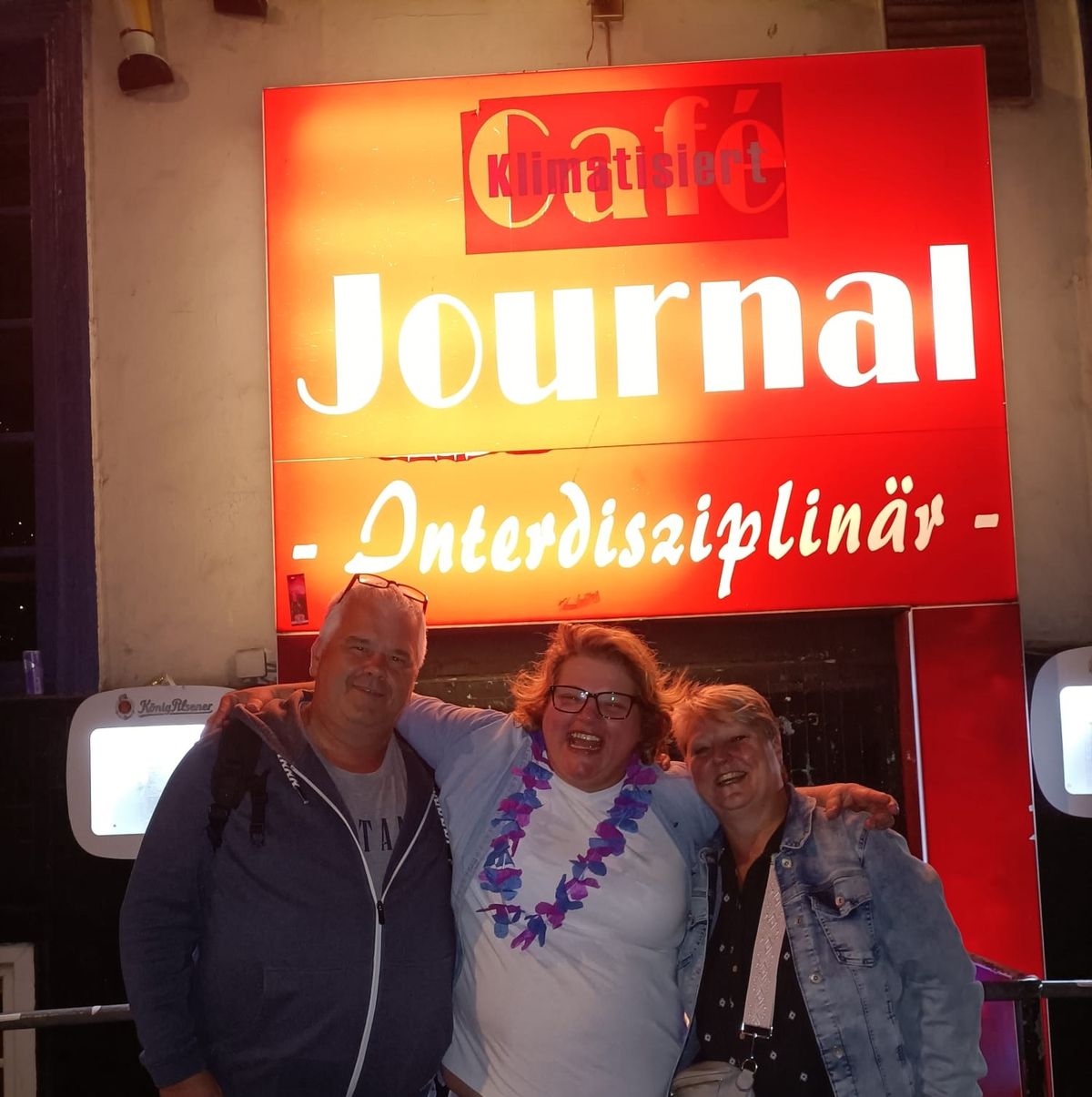 Drei lachende Personen vor dem leuchtenden Journal-Schild